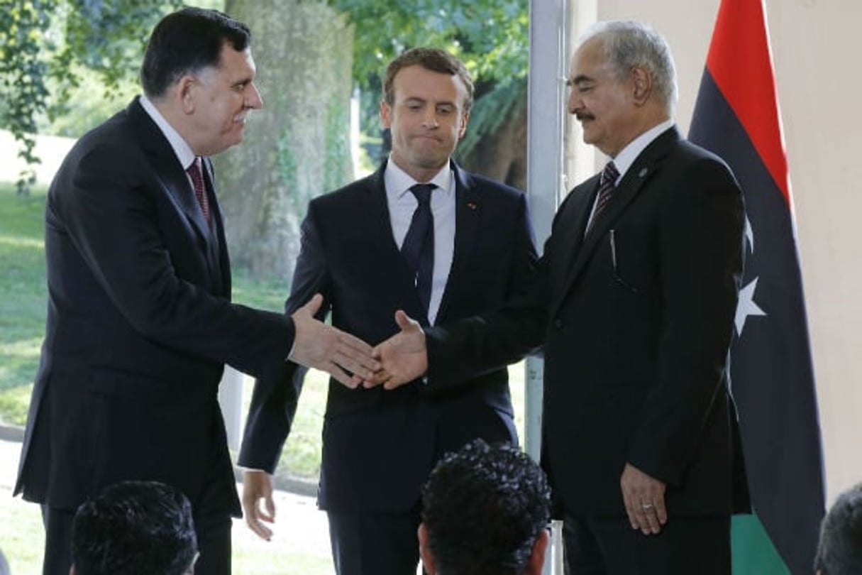 Le président du conseil présidentiel libyen Fayez al-Sarraj (g.) et le commandant de l’armée nationale libyenne Khalifa Haftar, se serrent la main sous le regard d’Emmanuel Macron, président français, à l’issue de la rencontre de ce mardi 25 juillet près de Paris. © Michel Euler/AP/SIPA