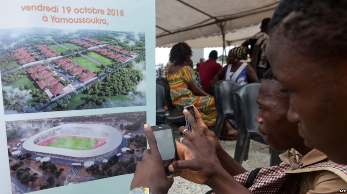 Les gens prennent des photos d’une pancarte représentant un futur stade à Yamoussoukro, le 19 octobre 2018. © AFP.
