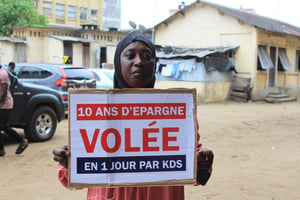 Une femme ayant investi dans KDS tient un panneau sur lequel est écrit « 10 ans d’épargne volée en 1 jour par KDS ». © DR.